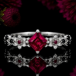 May include: A silver ring featuring a central princess-cut deep red gemstone. The band is decorated with two silver floral motifs, each centered with a small red stone, flanked by small black stones set into the metal along the band's sides.