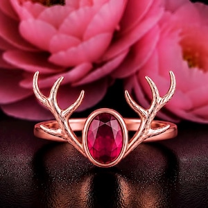 Peut inclure: Une bague en or rose ornée d'une pierre précieuse rouge foncé de taille ovale au centre. L'anneau se prolonge par deux branches symétriques et détaillées en forme de bois de cerf qui encadrent la pierre, offrant une finition métallique polie.
