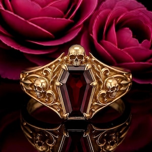 Peut inclure: Une bague en or ornée d'une pierre précieuse rouge foncé en forme de cercueil. L'anneau présente des volutes complexes et trois petits crânes, l'un placé au-dessus du cercueil et deux sur les côtés de la monture en métal.