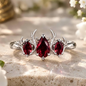 Puede incluir: Un anillo de tono plateado con tres piedras preciosas de color rojo intenso en forma de pera dispuestas en fila. La piedra central es la más grande, flanqueada por dos más pequeñas. Cada piedra está engastada con garras metálicas ramificadas.