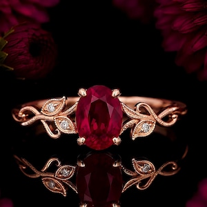 May include: A rose gold ring featuring a central oval-cut deep red ruby held by four prongs. The band is designed with intricate metal leaf motifs on both sides, each accented with a small, round, clear white diamond set within the foliage.
