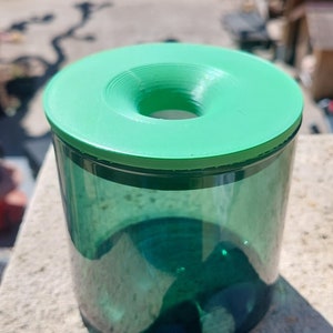Peut inclure: Un pot en verre vert avec un couvercle vert et un trou au centre du couvercle. Le pot est posé sur une surface gris clair.
