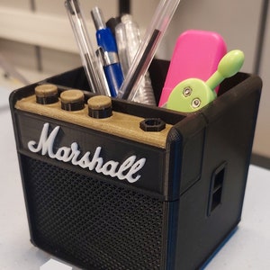 Peut inclure: Un organisateur de bureau imprimé en 3D noir et or en forme d'ampli Marshall. L'organisateur a une grille de haut-parleur noire et un dessus doré avec le mot "Marshall" imprimé en blanc. L'organisateur est rempli de stylos, de crayons et d'autres fournitures de bureau.