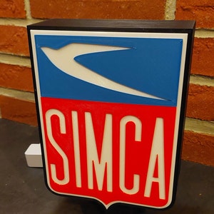 Può includere: Un cartello stampato in 3D in bianco e nero con un logo blu e bianco e la parola "SIMCA" in lettere bianche su sfondo rosso.