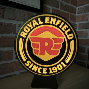 May include: A black and yellow round sign with the words "Royal Enfield" and "Since 1901" around a red and yellow logo with the letter "R" in the centre. The sign is on a black stand.