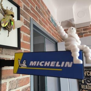May include: A Michelin sign with the Michelin Man figure sits on a brick wall. The sign is blue and yellow with white text. A framed insect artwork is also visible. The scene is indoors.