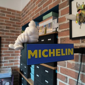 Peut inclure: Une figurine blanche de Bibendum Michelin est assise au-dessus d'une enseigne bleue avec le texte jaune "MICHELIN". L'enseigne est montée sur une structure noire. L'arrière-plan présente un mur de briques et des étagères.