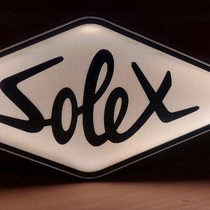 May include: A white diamond-shaped sign with a black border and the word "Solex" written in a stylised black font.