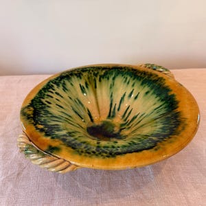 May include: A round ceramic bowl featuring a glossy glaze with a gradient of mustard yellow at the rim transitioning into a deep forest green center with radiating brushstroke patterns. The bowl includes two decorative twisted rope-style handles.