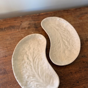 Puede incluir: Dos platos de cerámica de color blanco roto, con forma de riñón y un diseño en relieve similar a una hoja. Los platos están colocados sobre una superficie de madera oscura, mostrando su forma y textura únicas. Ideales para aperitivos.