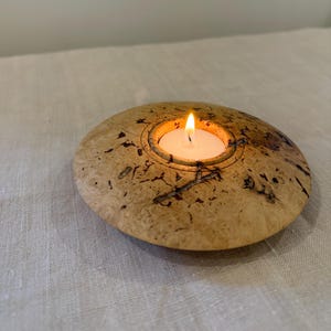 Puede incluir: Un portavelas redondo de madera con una vela blanca encendida. El portavelas está hecho de madera de color claro con marcas marrón oscuro y negro. La llama de la vela es brillante y el soporte está sobre una superficie neutra.