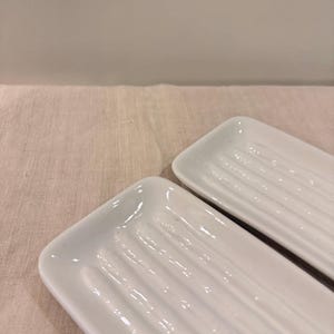 May include: Two rectangular, white ceramic soap dishes with a ridged design. The dishes have rounded corners and a glossy finish. The background is a neutral color.