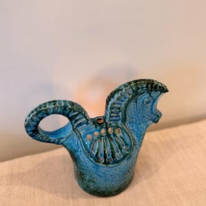 Puede incluir: Jarra de cerámica turquesa con forma de cabeza de caballo. La jarra tiene un asa y detalles decorativos, incluyendo una superficie texturizada y pequeñas aberturas circulares. La cabeza del caballo tiene rasgos tallados y una melena marrón oscuro.