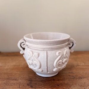 Może przedstawiać: Kremowy ceramiczny pojemnik na rośliny z dwoma uchwytami i dekoracyjnymi detalami reliefowymi. Pojemnik ma zaokrąglony kształt z pionowymi liniami i ornamentami. Neutralny kolor i klasyczny design sprawiają, że pasuje do różnych stylów wystroju.