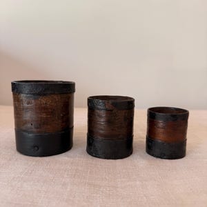 Puede incluir: Tres recipientes cilíndricos de diferentes tamaños, cada uno con un cuerpo de grano de madera marrón oscuro acentuado por una banda negra sólida en la base y un borde negro a juego en la parte superior. Los recipientes están dispuestos de mayor a menor.