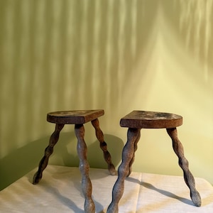 May include: Two antique wooden stools. Each has a triangular seat and three twisted legs. The stools are made of weathered wood with a natural finish. They are placed on a white surface against a light green wall.