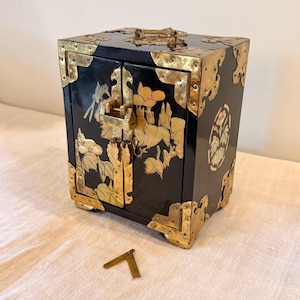 May include: Black wooden cabinet with gold-colored metal accents and floral designs. The cabinet has two doors, a lock, and decorative corner pieces. A small, gold-colored metal piece lies in front of the cabinet.