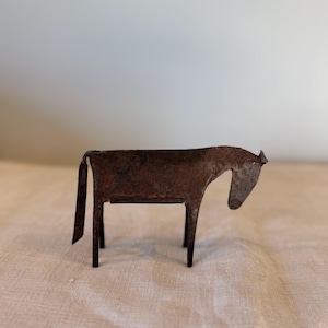 May include: A small, rustic metal horse sculpture with a weathered, brown finish. The horse has a simple, geometric design with a rectangular body, four legs, and a curved neck. The sculpture is approximately 13 cm long.