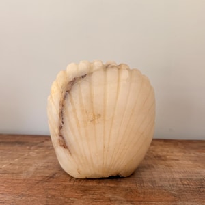 May include: A carved stone sculpture shaped like a scallop shell, featuring a translucent off-white and cream colour palette with natural brown veining running vertically along the textured, ribbed surface of the curved decorative object.