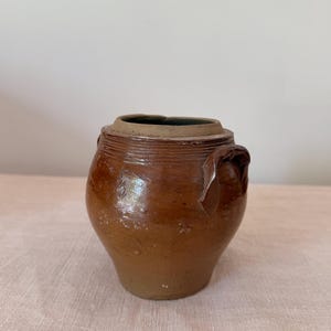 May include: A brown glazed ceramic jar with a wide mouth, a thick rim, and two small side handles. The exterior surface features horizontal ridges near the top opening and a smooth, rounded body that tapers toward a narrow, flat base.