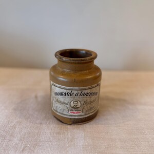 May include: A small, antique mustard jar with a tan exterior and a label featuring the text "moutarde à l'ancienne Armand Bizouard". The label also includes a portrait and the word "AMORA". The jar has a rounded body and a narrow opening.