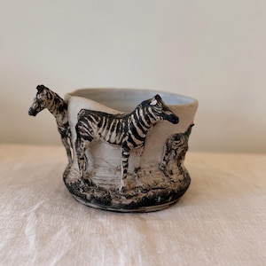 以下が含まれることがあります： オフホワイトの質感のある外観のセラミックボウル。ボウルの外側には、白黒の立体的なシマウマのフィギュアが施されています。ベースとリムは黒く塗装されており、ボウルの明るいボディとのコントラストを生み出しています。