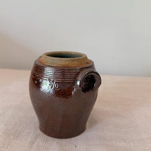 May include: A small, dark brown glazed stoneware crock featuring a rounded body, a wide circular opening with a thick rim, and a single side handle. The exterior surface displays a glossy finish with horizontal ridges and the embossed number 1/2.