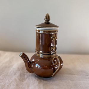 May include: A brown ceramic teapot with a unique design. The teapot has a bulbous base and a tall, cylindrical top with a lid. The spout and handle are curved, and the overall finish is glossy.
