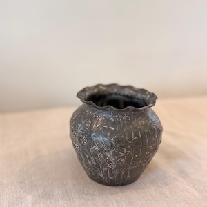 May include: A dark gray metal vase with a ruffled rim. The vase has an embossed floral design. The vase is sitting on a light-colored surface. The vase is likely used for decorative purposes.