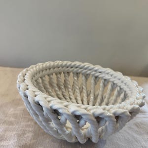 Puede incluir: Una cesta de cerámica blanca con un diseño de cuerda trenzada. La cesta de forma ovalada tiene un acabado brillante y está sobre una superficie de color neutro. El tejido abierto de la cesta permite ver el contenido.