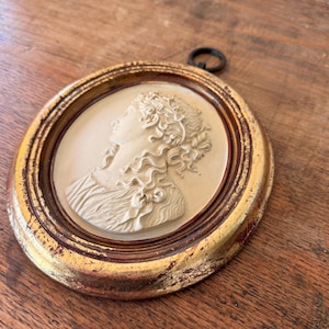 May include: Oval-shaped antique wall decor featuring a cream-colored cameo of a woman in profile. The cameo is set within a gold-painted, distressed wooden frame. A small, black metal ring is attached at the top for hanging.