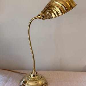 Puede incluir: Lámpara de escritorio vintage de latón con cuello de cisne flexible y pantalla en forma de concha. La lámpara tiene una base redonda y un acabado pulido, adecuada para una oficina en casa o estudio. La lámpara mide unos 38 cm de alto.