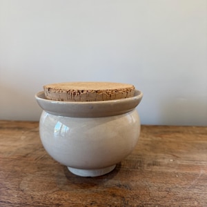 May include: A small, cream-coloured ceramic jar with a rounded body and a cork stopper. The jar has a wide, flat rim and sits on a small, circular base. The cork stopper is light brown and fits snugly into the jar's opening. The jar is on a wooden surface.