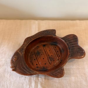 Puede incluir: Un jabonera de cerámica marrón con forma de pez. El plato tiene una depresión central con agujeros de drenaje y detalles grabados. Un accesorio de baño decorativo y funcional.