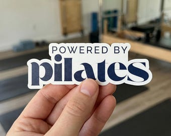 Può includere: Un adesivo bianco con la scritta "POWERED BY pilates" in blu navy, con un carattere stilizzato. L'adesivo è tenuto in mano, con uno studio di pilates sullo sfondo.
