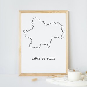 May include: A framed print featuring a minimalist outline of the Saône-et-Loire region in France, rendered in black on a white background. The text "SAÔNE ET LOIRE" is printed below the map. A candle and decorative elements are in the foreground.
