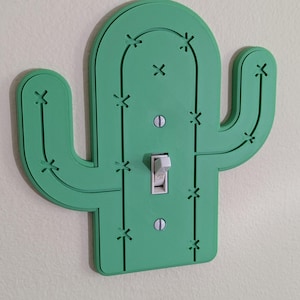 May include: A green cactus-shaped light switch cover with a white toggle switch. The cover has decorative cutouts and is mounted on a light-colored wall. The switch is in the "ON" position.