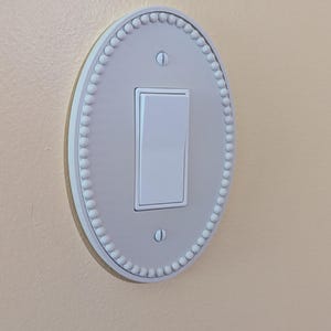 May include: A white light switch with an oval, beaded plate, mounted on a light beige wall. The switch is in the 'on' position. The plate is a decorative feature.