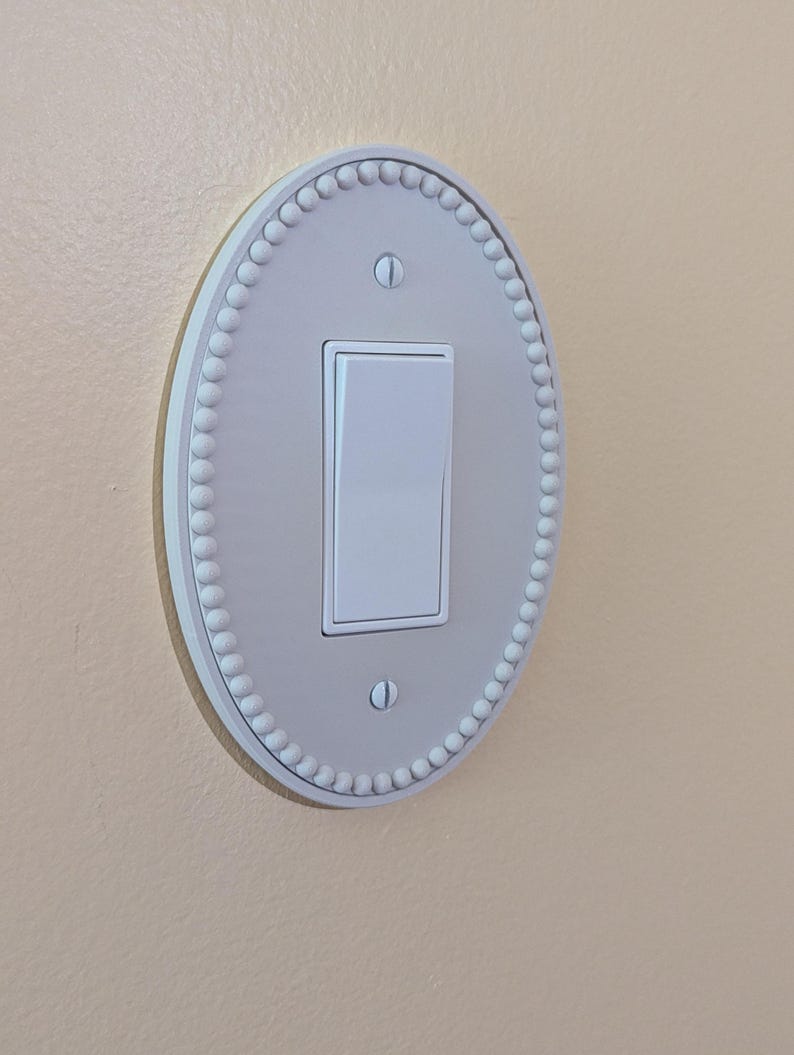 May include: A white light switch with an oval, beaded plate, mounted on a light beige wall. The switch is in the 'on' position. The plate is a decorative feature.