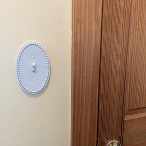 May include: A pale blue, oval light switch cover with a beaded edge. The switch is flipped upwards. The cover is fixed to a cream wall, adjacent to a wooden door with a brass handle.