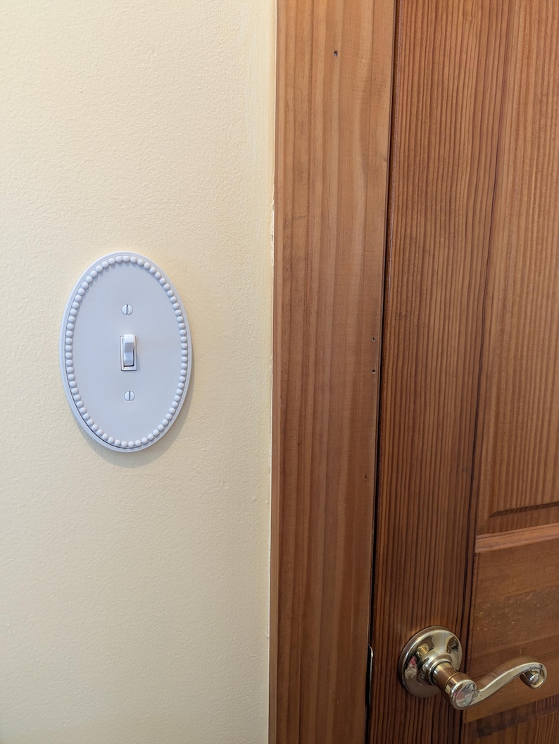 May include: A pale blue, oval light switch cover with a beaded edge. The switch is flipped upwards. The cover is fixed to a cream wall, adjacent to a wooden door with a brass handle.