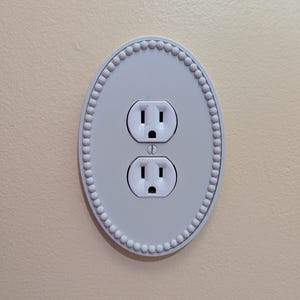May include: A white, oval-shaped electrical outlet cover with a beaded border. The cover has two standard electrical outlets. The wall is painted a light beige colour.