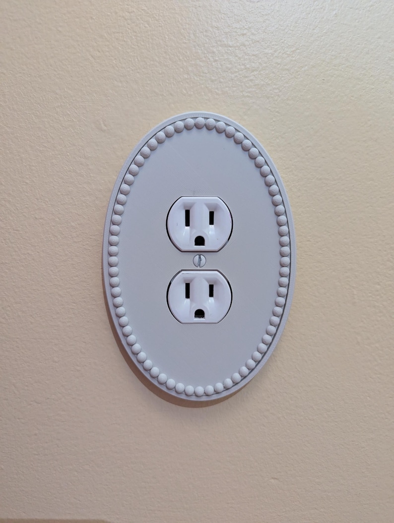 May include: A white, oval-shaped electrical outlet cover with a beaded border. The cover has two standard electrical outlets. The wall is painted a light beige colour.