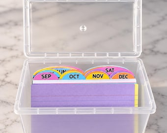 May include: A clear plastic storage box with a hinged lid containing a set of colorful index cards. The cards feature tabbed dividers labeled with months including SEP, OCT, NOV, and DEC, along with days of the week like SAT, in various bright colors.
