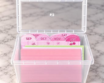 May include: A clear plastic storage box with an open hinged lid containing a set of colorful index cards. The cards feature pink, green, and white dividers labeled with months including SEP, OCT, NOV, and DEC, along with days like SUN and SAT.