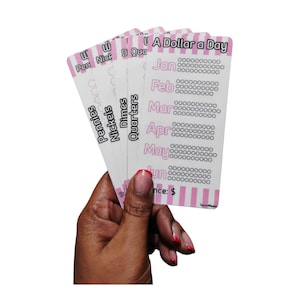 Puede incluir: Una mano sostiene una pila de tarjetas rosas y blancas. Las tarjetas están etiquetadas con las palabras "Pennies", "Nickels", "Dimes" y "Quarters". Las tarjetas también tienen el texto "A Dollar a Day" y los meses del año.