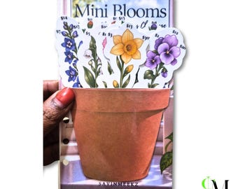 May include: A collection of die-cut flower stickers featuring blue delphiniums, white lily of the valley, a yellow daffodil, and purple pansies, arranged in a terracotta-colored pot illustration on a card labeled Mini Blooms.