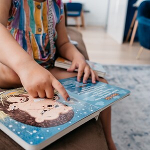 May include: A child sits on a couch and points at a book with a blue cover and a cartoon illustration of a hedgehog. The book has the words "Sleepy Time" printed on the cover.