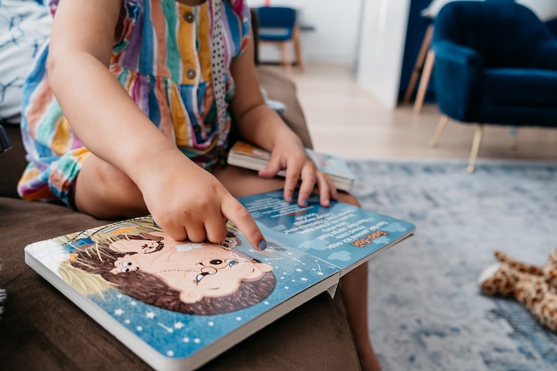 May include: A child sits on a couch and points at a book with a blue cover and a cartoon illustration of a hedgehog. The book has the words "Sleepy Time" printed on the cover.