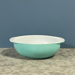 May include: A teal and white mixing bowl. The bowl has a rounded shape with a white interior and a teal exterior. The rim of the bowl is white. The bowl is sitting on a textured surface.